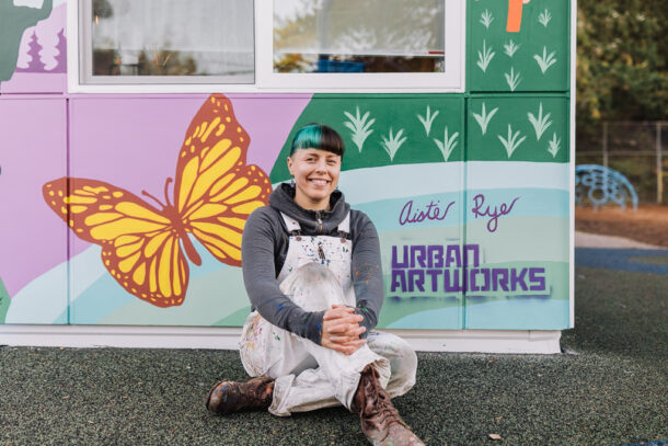 Artist Aistė Rye sitting in front of a mural she designed for the Northwest School for Deaf and Hard-of-Hearing Children. she is sitting in front of a butterfly her artist's signature and the Urban ArtWorks tag.